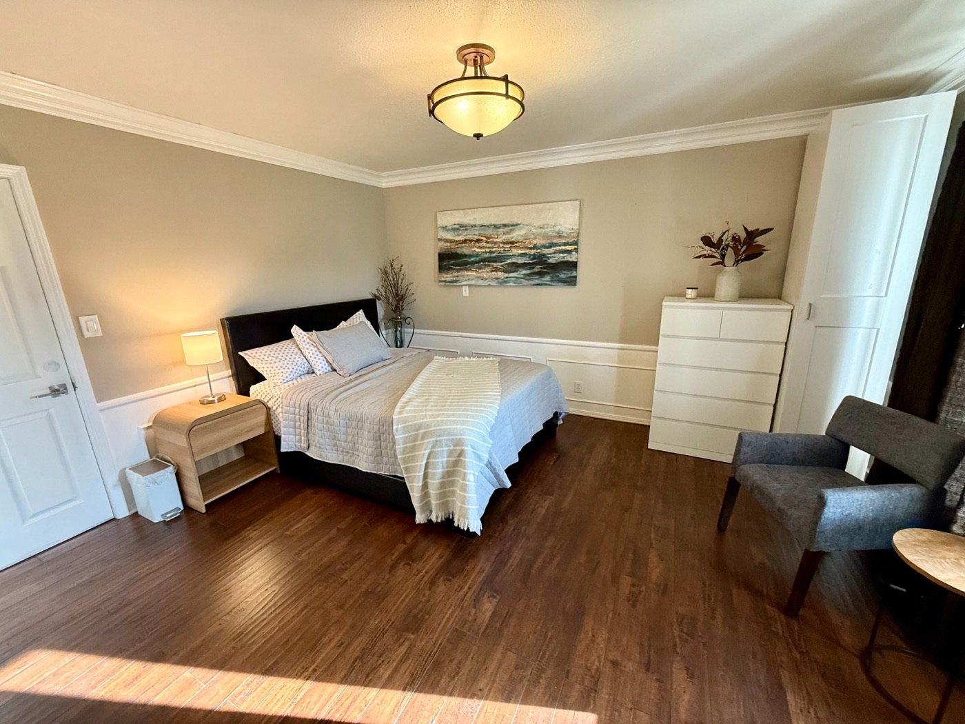 A furnished resident bedroom at Loving Hands with a made bed, dresser, armchair and wood floors